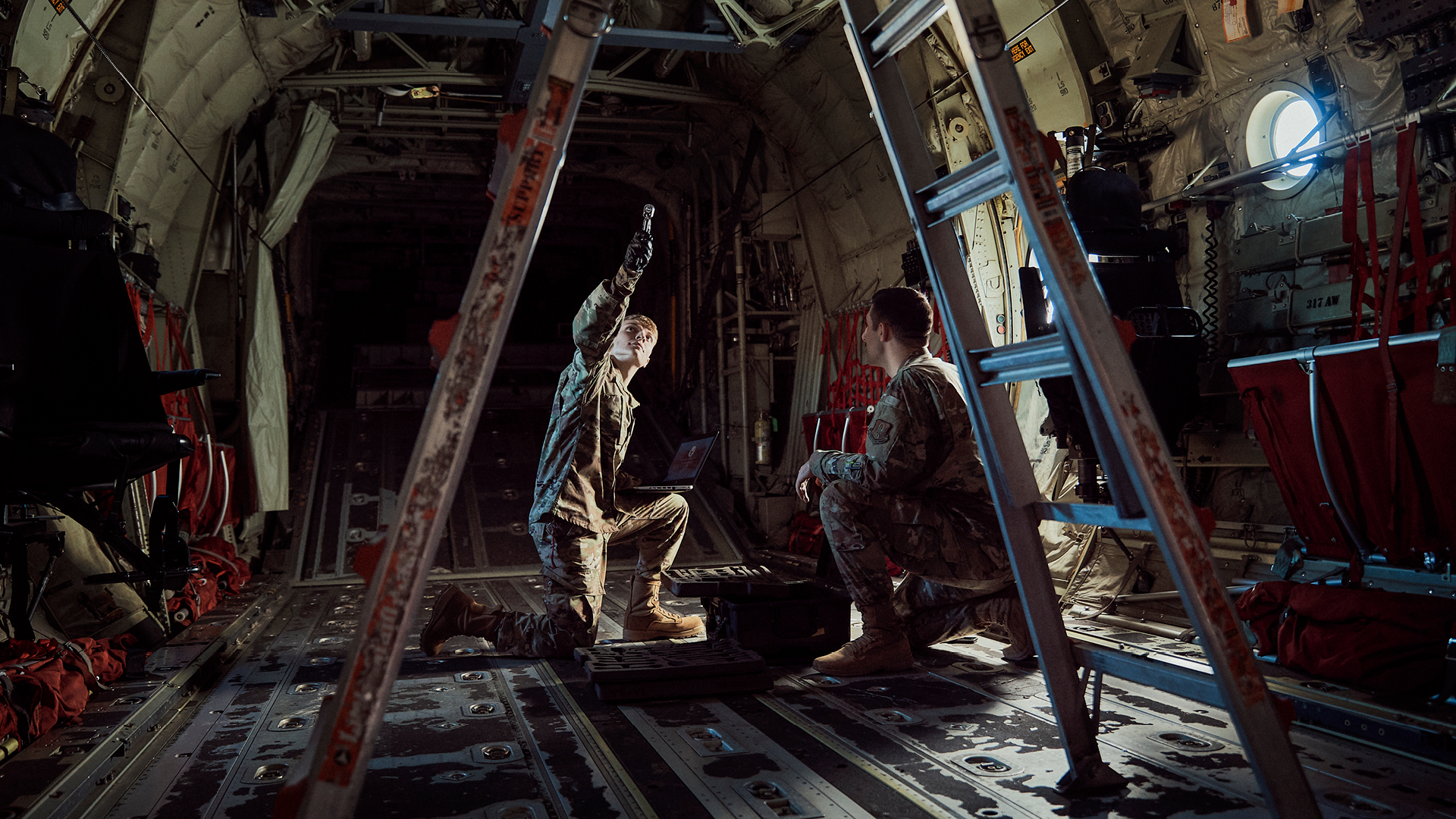Corbin Farrar pointing towards the ceiling of a plane while talking with a coworker
